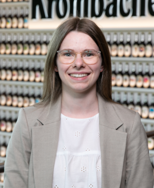Frau mit langen, blonden Haaren trägt eine graue Jacke und eine Brille. Sie lächelt freundlich in die Kamera.