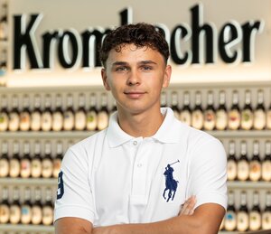 Man mit kurzen gelockten, braunen, Haaren trägt ein weißes T-Shirt und lächelt in die Kamera.