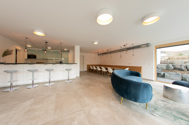 Moderne Innenansicht eines Raumes mit einer Küchenzeile, Barhockern, einem blauen Sofa und einem langen Esstisch mit weißen Stühlen.