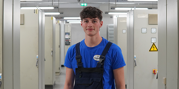 Junger Mann mit kurzen braun-gelockten Haaren trägt ein blaues T-Shirt, dazu eine dunkelblaue Latzhose. 