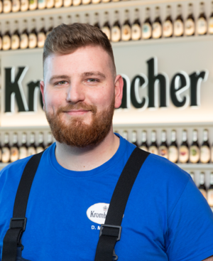 Mann mit kurzen, rötlichen Haaren trägt ein blaues T-Shirt mit Krombacher Logo Aufdruck. Er lächelt in die Kamera.