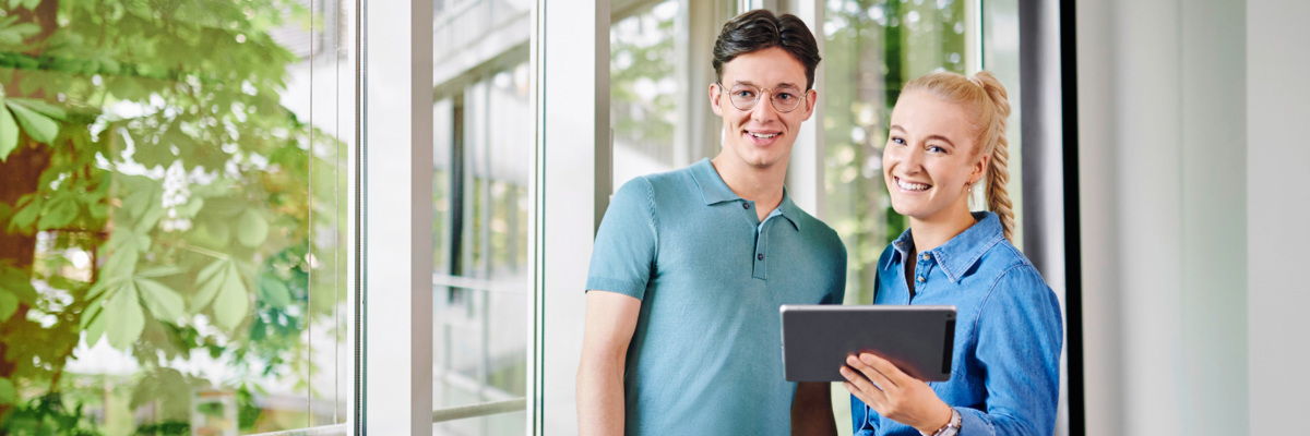Ein Mann in einem hellblauen Polohemd und eine Frau in einer blauen Jeansjacke stehen nebeneinander. Die Frau hält ein Tablet in der Hand. Im Hintergrund sind große Fenster mit Pflanzen zu sehen.