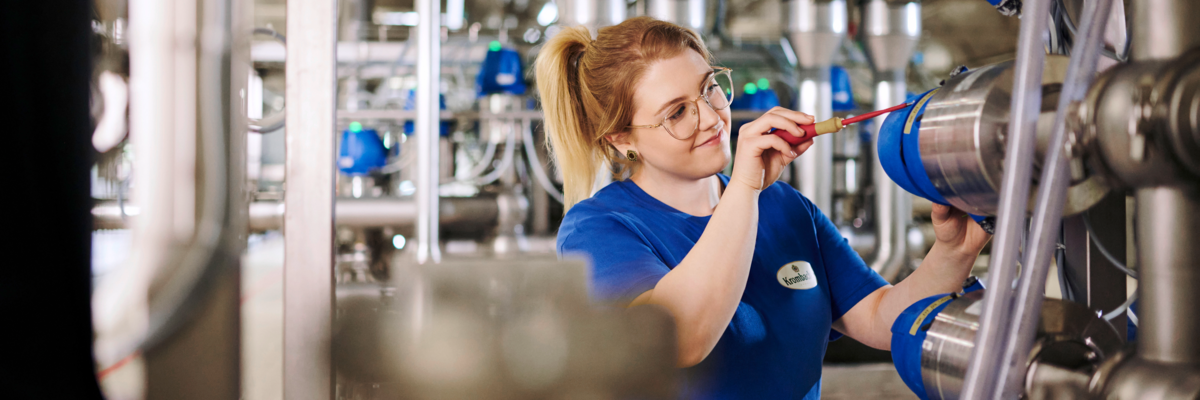Eine junge Frau in blauer Arbeitskleidung arbeitet an einer Maschine mit mehreren blauen Rohren und Metallleitungen. Werkzeuge sind sichtbar.