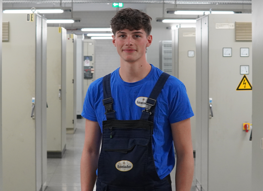 Junger Mann mit kurzen braun-gelockten Haaren trägt ein blaues T-Shirt, dazu eine dunkelblaue Latzhose. 