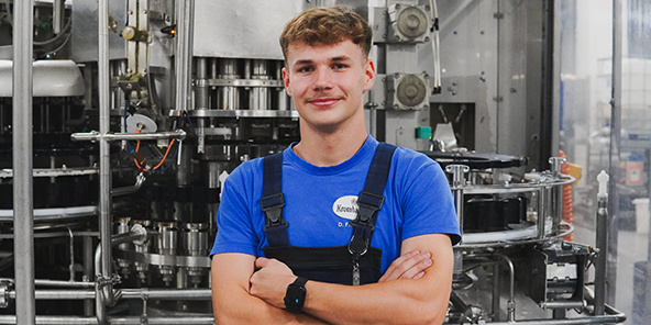 Mann mit dunkelblonden gelockten Haaren, trägt ein blaues T-Shirt und dunkelblaue Latzhose.