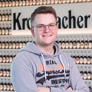 Mann mit dunkelblonden Haaren, trägt eine Brille und einen grauen Kapuzenpullover. Er lächelt in die Kamera.