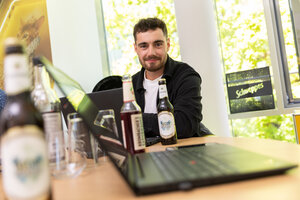 Ein Mann mit dunkel gelocktem Haar, sitzt an einem Tisch mit einem Laptop, er trägt ein dunkles Hemd.