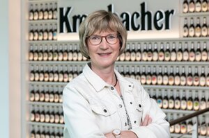 Frau mit blonden Haaren und Bob, trägt eine Brille und weiße Jeansjacke. Sie lächelt in die Kamera.
