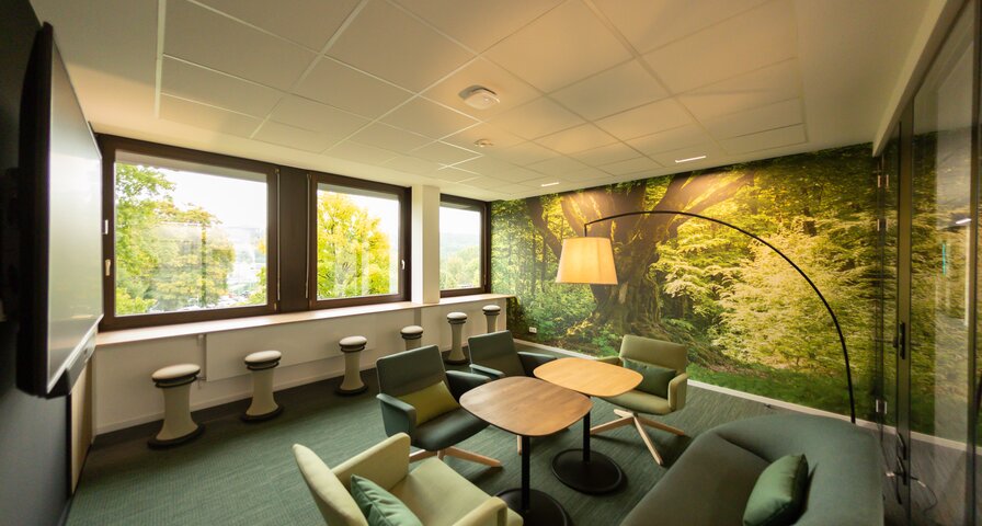 In grün gestalteter Besprechungsraum mit Sitzmöglichkeiten, einer Fototapete in Waldoptik und einer in den Raum hineinragender Stehlampe. 