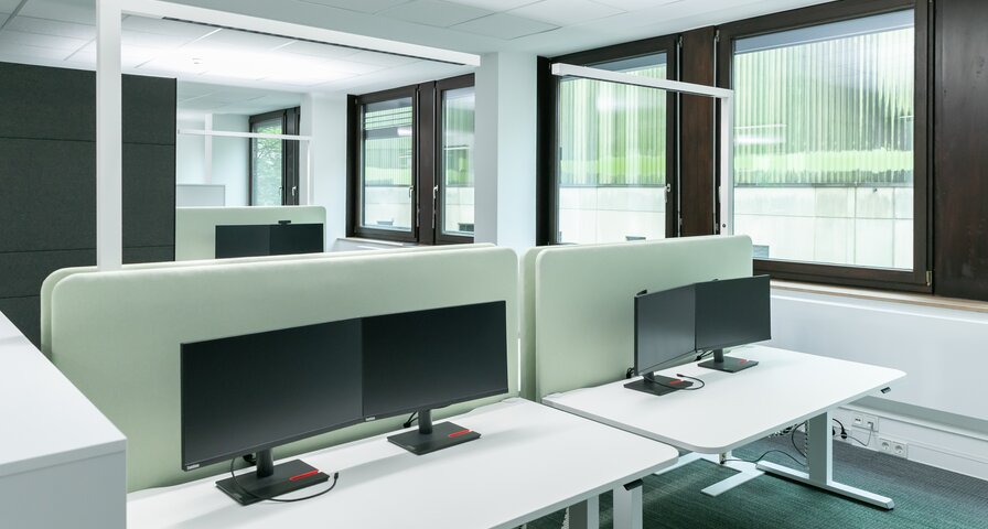 Büro mit zwei nebeneinander stehenden Schreibtischen, mit jeweils zwei Monitoren. Dahinter eine hellgrüne Trennwand.