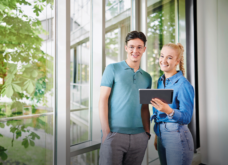 Ein Mann in einem hellblauen Polohemd und eine Frau in einer blauen Jeansjacke stehen nebeneinander. Die Frau hält ein Tablet in der Hand. Im Hintergrund sind große Fenster mit Pflanzen zu sehen.