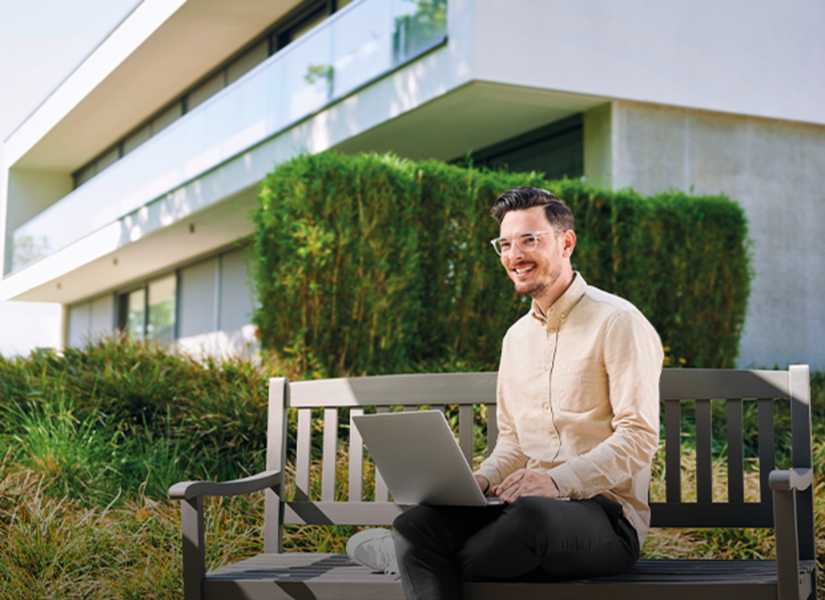 Mann sitzt auf einer Bank im Freien und arbeitet an einem Laptop. Im Hintergrund sind moderne Gebäude und grüne Sträucher sichtbar.