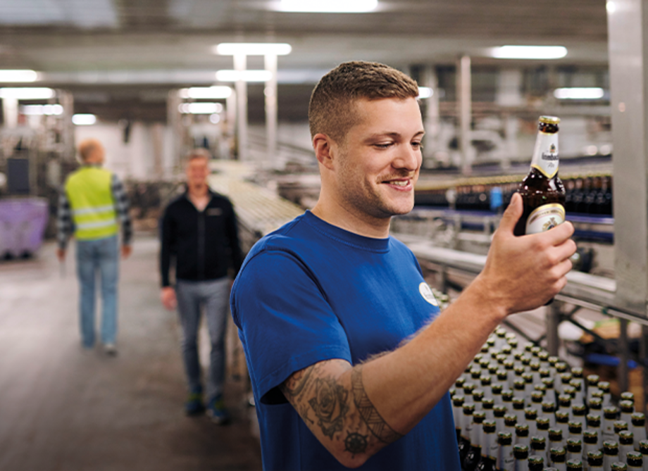 Mann mit tätowiertem Arm hält eine Flasche Bier in der Hand. Im Hintergrund sind weitere Personen und Flaschen auf einem Förderband zu sehen.