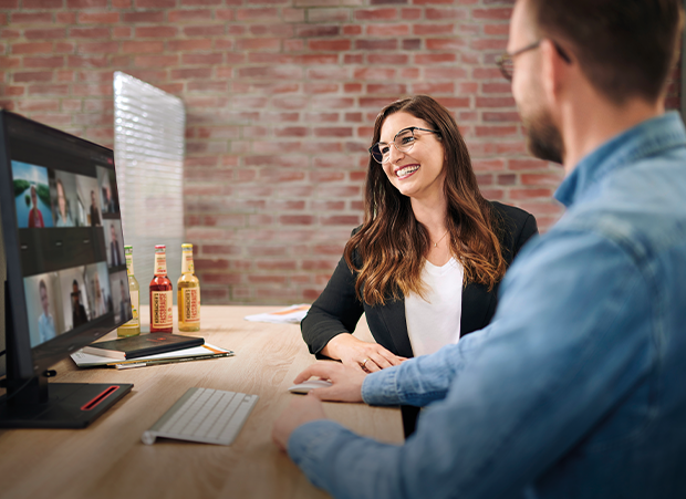 Eine Frau mit langen, braunen Haaren in einem schwarzen Blazer sitzt an einem Tisch und schaut auf einen Laptop. Auf dem Tisch stehen zwei Flaschen und eine Tastatur.