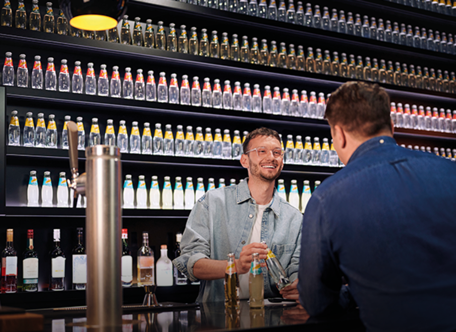 Zwei Personen stehen an einer Bar. Eine Person trägt eine Jeansjacke und hält eine Schweppes Flasche, während die andere Person vor der Bar sitzt.