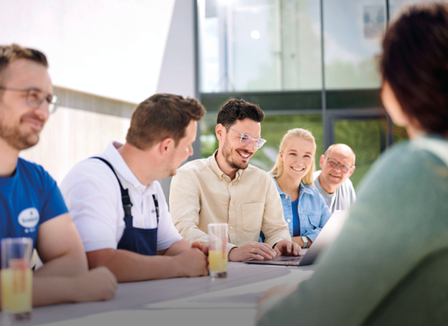 Fünf Personen sitzen an einem Tisch in einem modernen Büro, diskutieren, lächeln und arbeiten gemeinsam an Laptops.