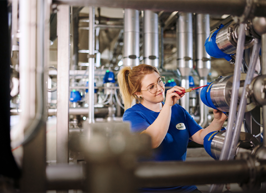 Eine junge Frau in blauer Arbeitskleidung arbeitet an einer Maschine mit mehreren blauen Rohren und Metallleitungen. Werkzeuge sind sichtbar.