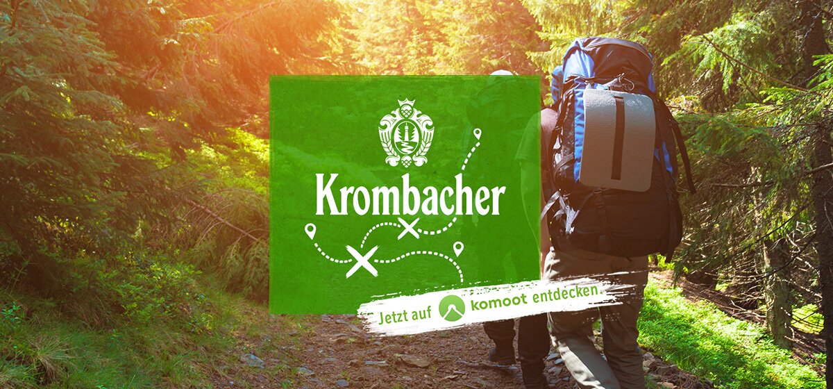 Zwei Wanderer mit Rucksäcken gehen einen Waldweg entlang. Im Vordergrund ist das grüne Logo von Krombacher mit dem Text 'Jetzt auf komoot entdecken'.