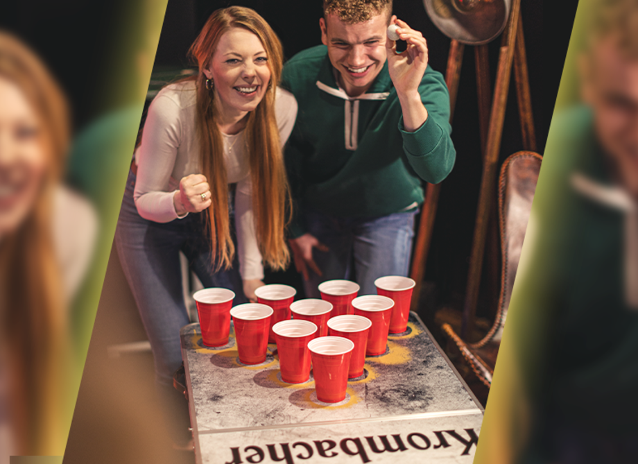 Zwei junge Menschen spielen Beerpong mit roten Plastikbechern auf einem Beerpong-Tisch mit dem Schriftzug 'Krombacher'
