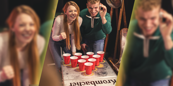 Zwei junge Menschen spielen Beerpong mit roten Plastikbechern auf einem Beerpong-Tisch mit dem Schriftzug 'Krombacher'