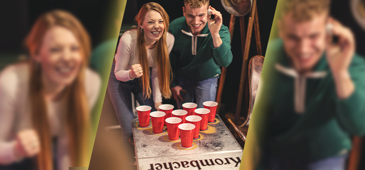Zwei junge Menschen spielen Beerpong mit roten Plastikbechern auf einem Beerpong-Tisch mit dem Schriftzug 'Krombacher'