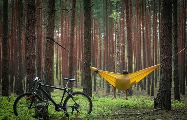 Eine gelbe Hängematte ist zwischen zwei Bäumen gespannt und jemand sitzt darin. Ein Fahrrad ist links neben der Hängematte an einen Baum gelehnt.