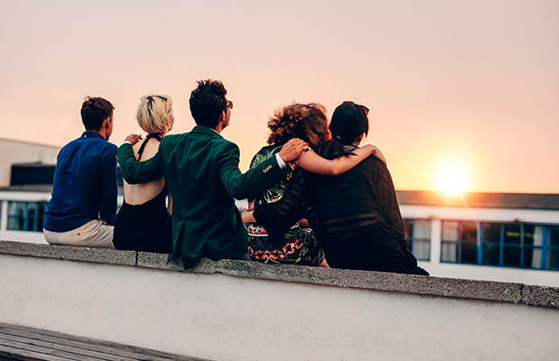 Fünf junge Leute sitzen mit dem Rücken zur Kamera auf einer Mauer und beobachten den Sonnenuntergang.