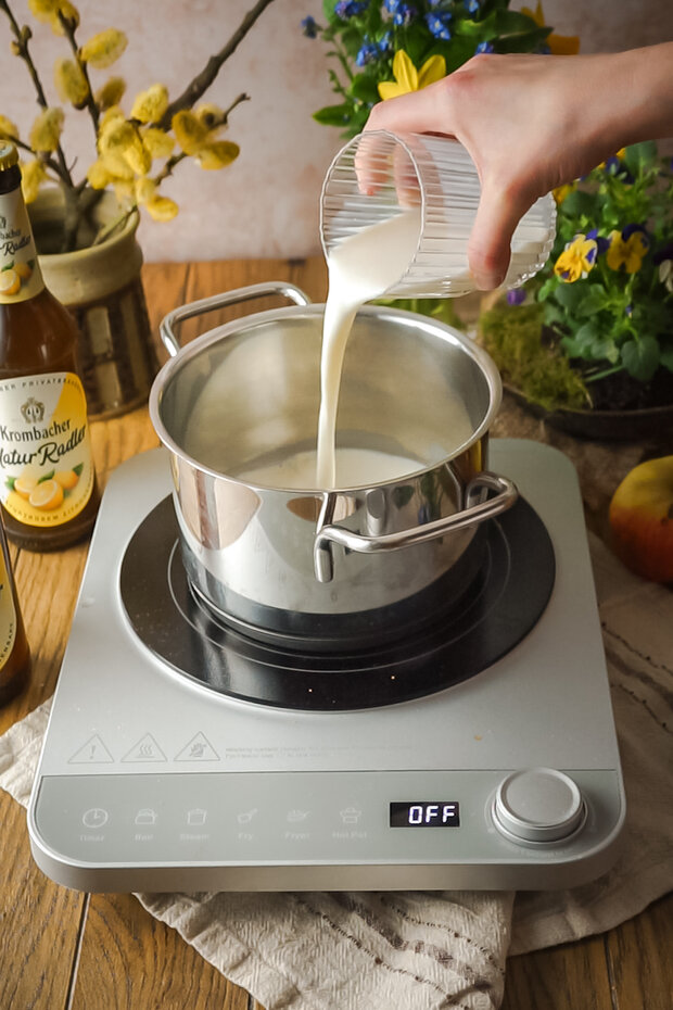Eine Hand gießt Milch aus einem transparenten Becher in einen silbernen Kochtopf auf einem elektrischen Kochfeld.