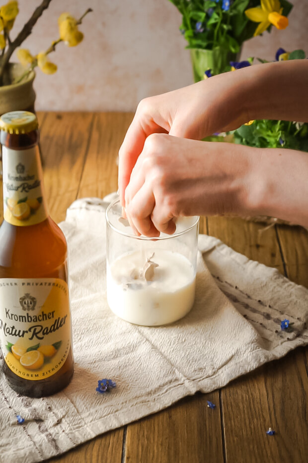 Eine Hand drückt Hefe in ein Glas mit einer hellen Flüssigkeit, daneben steht eine Flasche Krombacher Natur Radler auf einem Tisch.