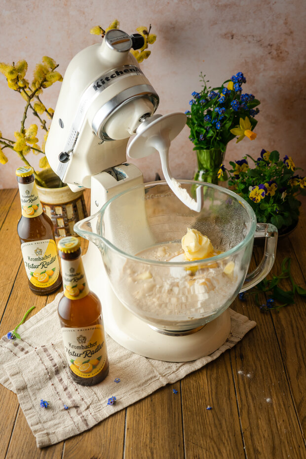 Eine Küchenmaschine mit einem transparenten Rührbehälter, in dem Teig mit Butter gemischt wird, umgeben von zwei Flaschen 'König Pilsener' und frischen Blumen.