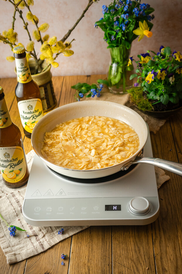 Eine Pfanne mit einer hellen Flüssigkeit und Mandeln auf einem Kochfeld, umgeben von zwei Flaschen 'Krombacher Natur Radler', einer Tischdecke und Blumen in einer Vase.