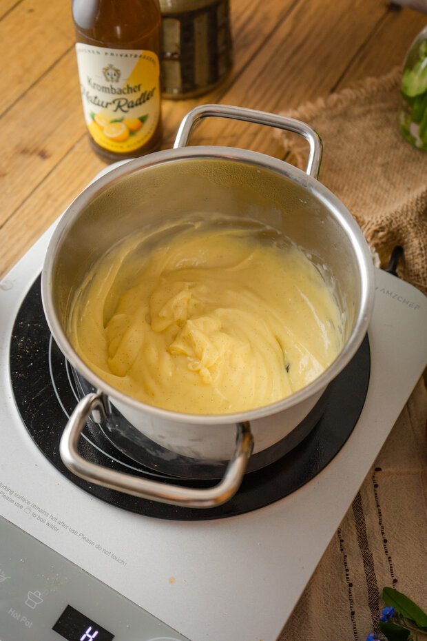Ein Topf mit cremiger, gelber Sauce steht auf einem Induktionskochfeld, im Hintergrund eine Flasche Kombacher mit Zitrone.