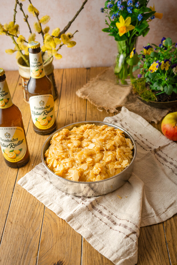 Eine Schüssel mit gelben Apfelstücken steht auf einem Holztisch, umgeben von zwei Flaschen 'Krombacher NaturRadler', einer Apfel und bunten Blumen in einer Vase.
