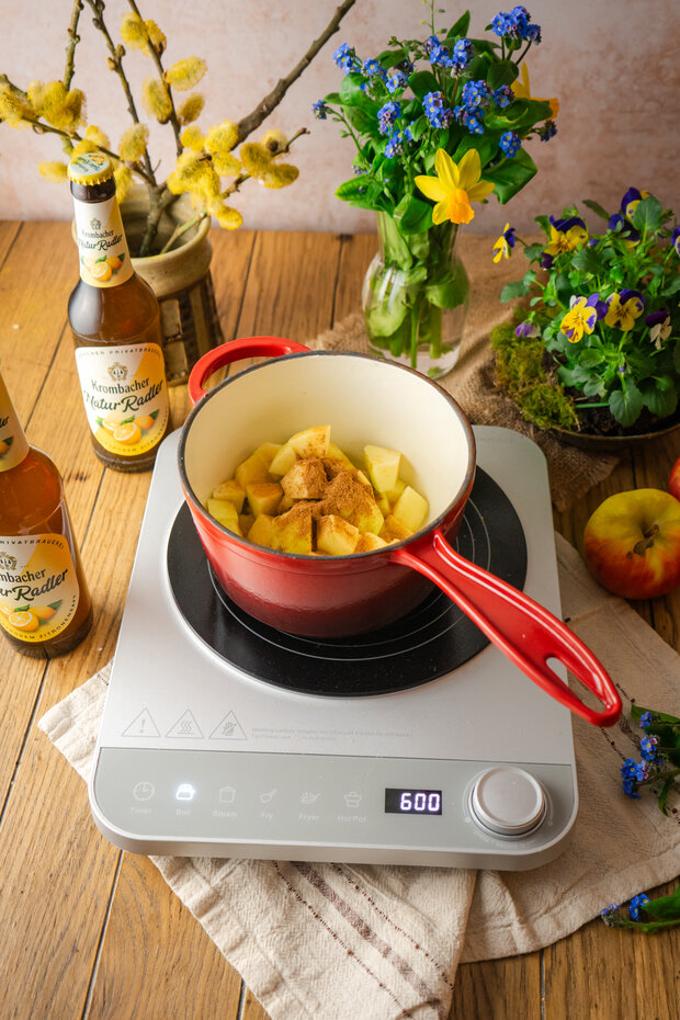 Roter Kochtopf mit Äpfeln und Gewürzen auf einem Induktionskochfeld, umgeben von Äpfeln und frischen Blumen.