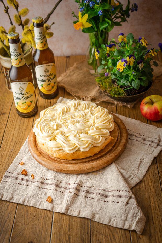 Ein runder Kuchen mit einer dekorativen Sahnehaube auf einem Holzbrett, umgeben von zwei Flaschen 'Kornbacher Natur Radler', einer Apfel und einer blühenden Pflanze.