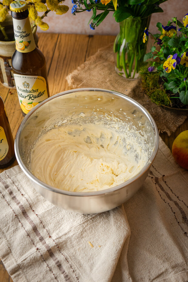 dEine silberne Schüssel mit cremiger, heller Masse steht auf einem Tisch, umgeben von einer Flasche 'Krombacher' und einer Flasche 'Natur-Radler', sowie einer Apfel und Blumen.