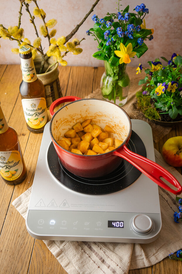 Roter Kochtopf mit Äpfeln und Gewürzen auf einem Induktionskochfeld, umgeben von zwei Bierflaschen, Äpfeln und frischen Blumen.