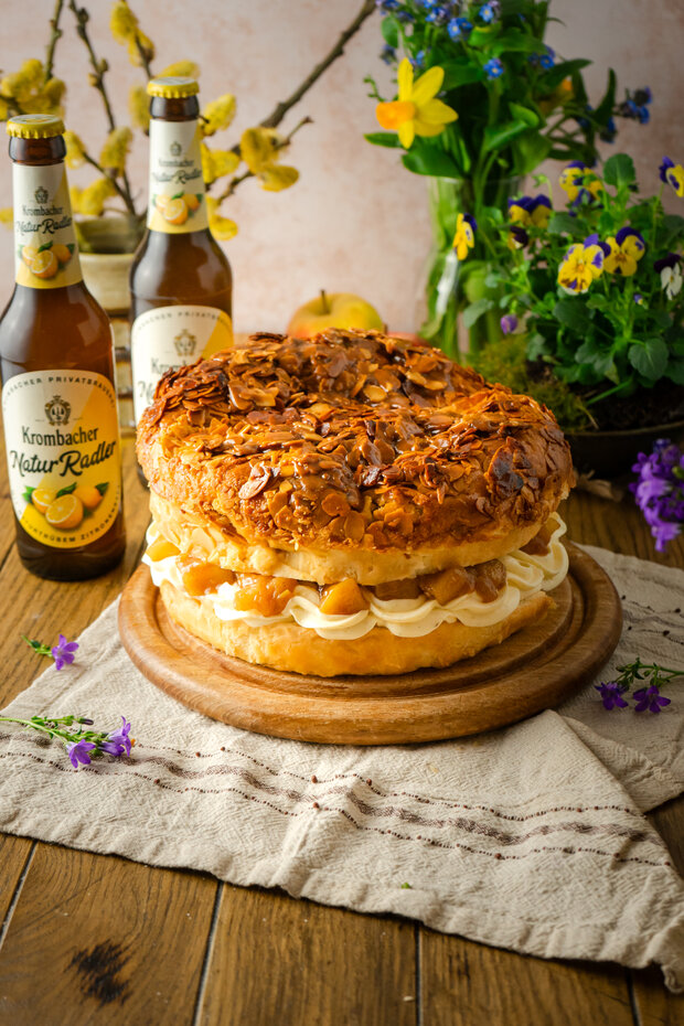 Ein großer, runder Kuchen mit einer knusprigen Mandelkruste, gefüllt mit Sahne und Früchten, steht auf einem Holzbrett, daneben zwei Flaschen Krombacher Natur-Radler.