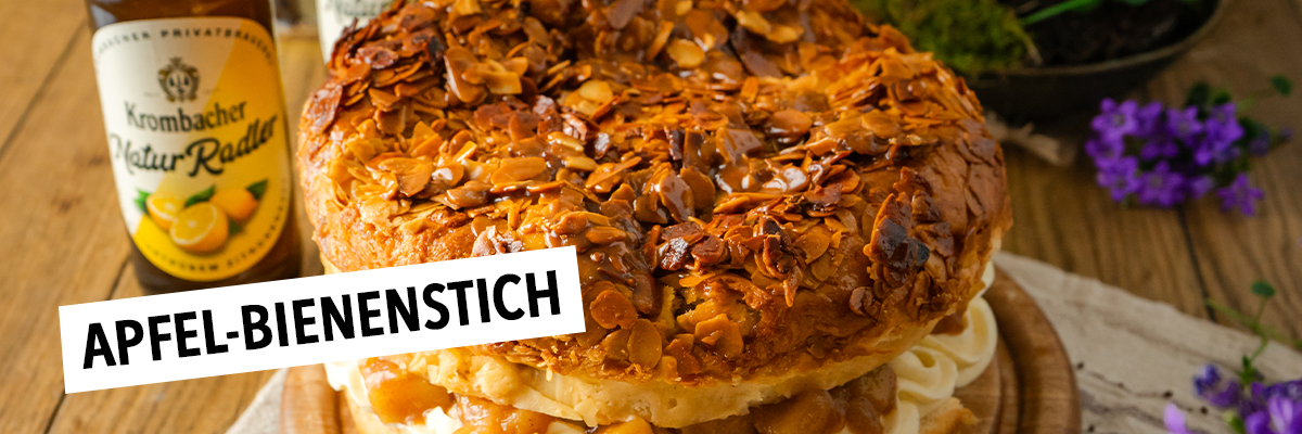 Ein großer, runder Kuchen mit einer knusprigen Mandelkruste, gefüllt mit Sahne und Früchten, steht auf einem Holzbrett, daneben zwei Flaschen Krombacher Natur-Radler.