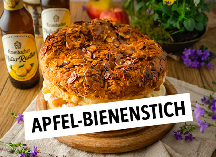 Ein großer, runder Kuchen mit einer knusprigen Mandelkruste, gefüllt mit Sahne und Früchten, steht auf einem Holzbrett, daneben zwei Flaschen Krombacher Natur-Radler.