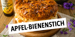 Ein großer, runder Kuchen mit einer knusprigen Mandelkruste, gefüllt mit Sahne und Früchten, steht auf einem Holzbrett, daneben zwei Flaschen Krombacher Natur-Radler.