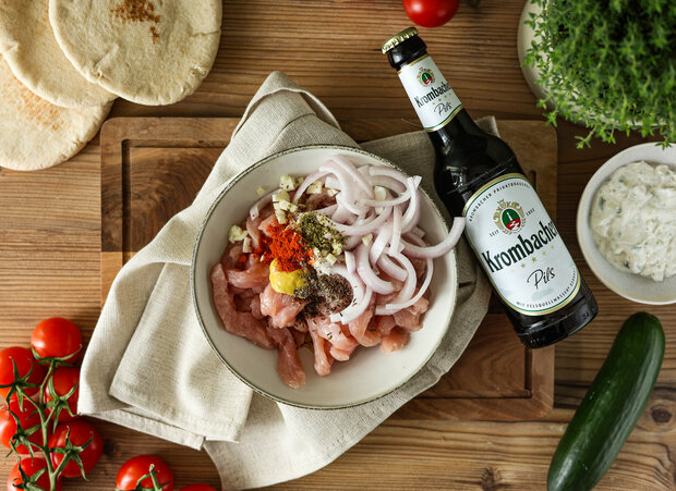 Eine Schüssel mit mariniertem Hähnchenfleisch, Zwiebeln und Gewürzen, daneben eine Flasche Kronbacher Pils, frische Tomaten, Gurke und Fladenbrot.