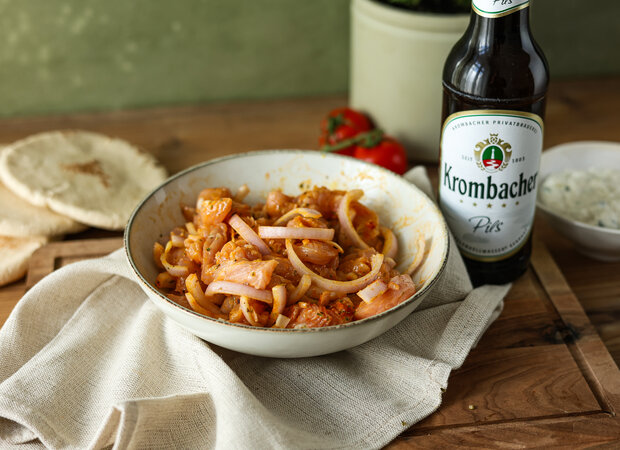 Eine Schüssel mit Fleisch, Zwiebeln und Tomatensauce, daneben ein Bier der Marke 'Krombacher' und Fladenbrot auf einem Holzuntergrund.