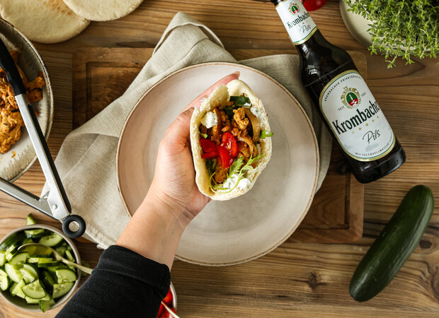 Eine Hand hält eine Tortilla mit Hähnchen, Salat und Tomaten, daneben eine Flasche Krombacher Pils und frische Zutaten auf einem Holztisch.