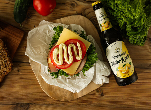 Ein Sandwich mit Käse, Schinken, Salat, Remoulade und Tomaten auf einem Holzbrettchen.
