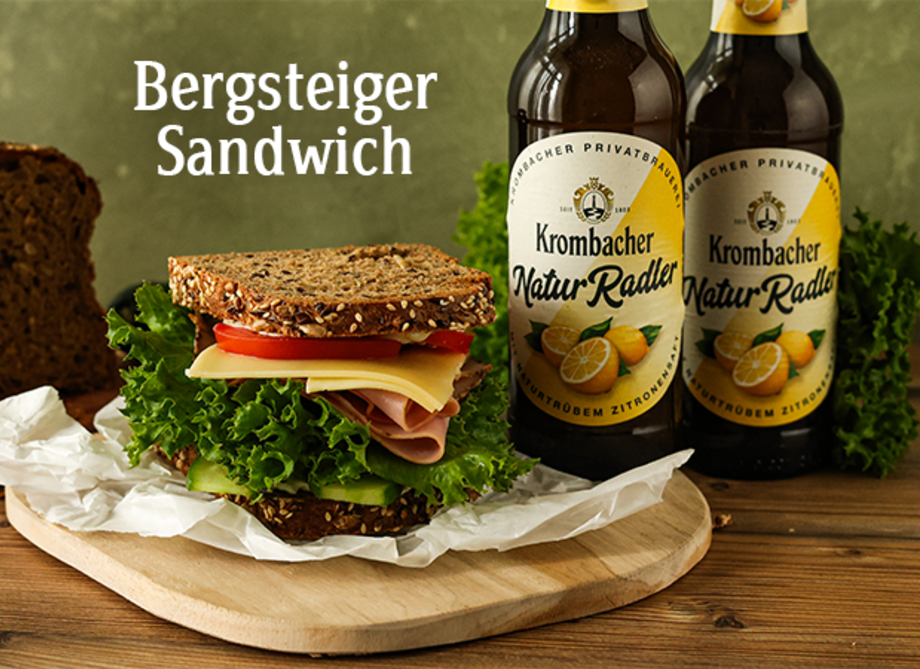 Ein Bergsteiger-Sandwich mit Käse, Schinken, Salat und Tomaten auf einem Holzbrettchen.