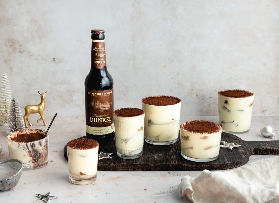 Sechs Gläser mit Tiramisu-Dessert stehen auf einem Holzbrett, einige mit Kakaopulver bestreut. Neben den Gläsern steht eine Flasche 'Dunkel' Bier.