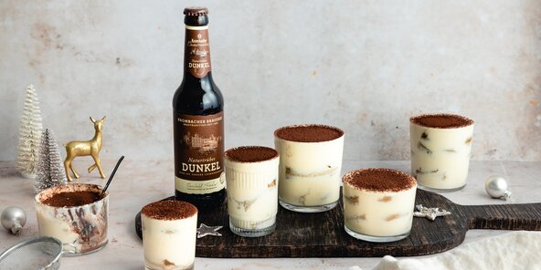 Sechs Gläser mit Tiramisu-Dessert stehen auf einem Holzbrett, einige mit Kakaopulver bestreut. Neben den Gläsern steht eine Flasche 'Dunkel' Bier.