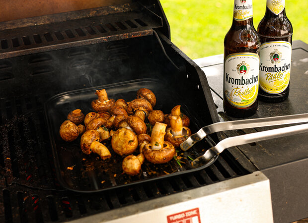 Eine Schale mit gegrillten Champignons auf einem Grill und neben Krombacher Radler.