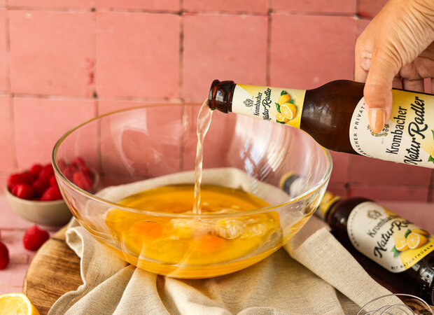 Eine Hand schüttet Krombacher Natur Radler in eine durchsichtige Schüssel mit mehreren Eigelben.